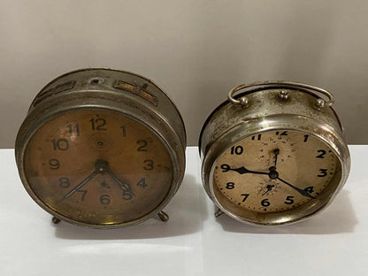 Two vintage silver-colored metal alarm clocks with worn faces on a white surface against a gray background